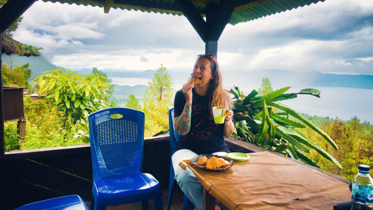 Carly (a blonde Wesetrn woman) sits eating a plate of fried bananas in a small gazebo with the backdrop of lake toba behind her