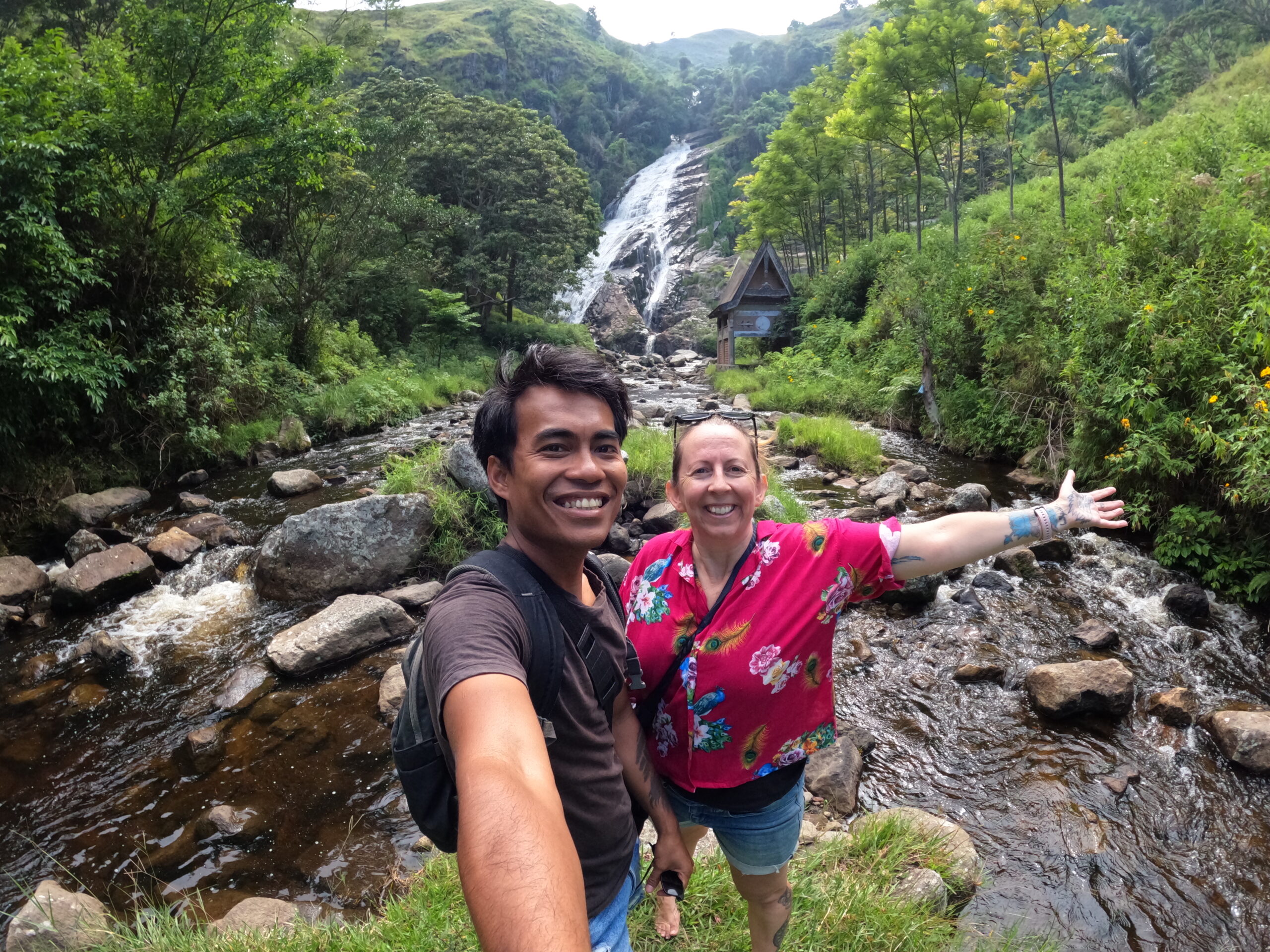 An Indonesian man and New Zealand woman stand with arms outstretched in front of a waterfall.